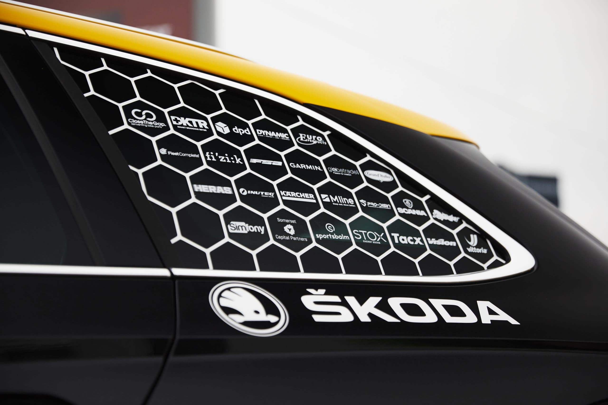 Zwarte en gele Škoda met sponsorbelettering op de zijruit, voorzien van witte honingraatprint, diverse logo’s en Škoda-voertuigreclame op de carrosserie.