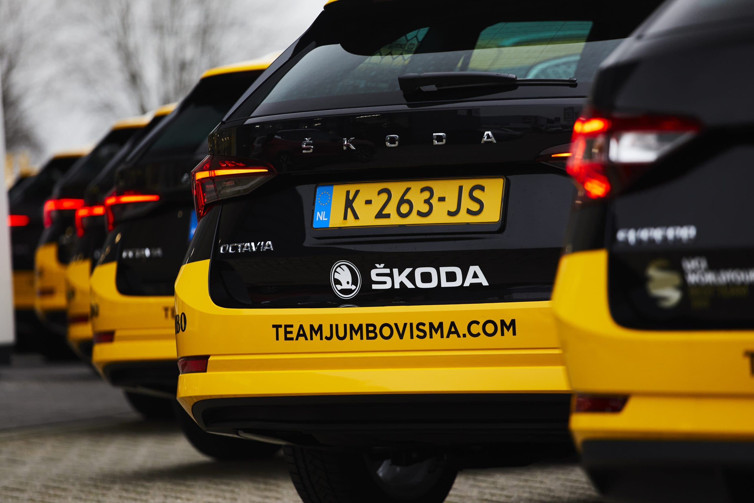 Rij zwarte en gele Škoda-teamauto’s met voertuigbelettering op de achterzijde, voorzien van Škoda-logo en websitebedrukking "teamjumbovisma.com" op de bumper.