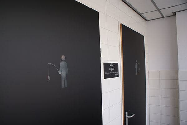 Zwarte toiletdeuren met subtiele sanitaire pictogrammen en wayfinding-bewegwijzering op een wit betegelde wand in een bedrijfsinterieur.