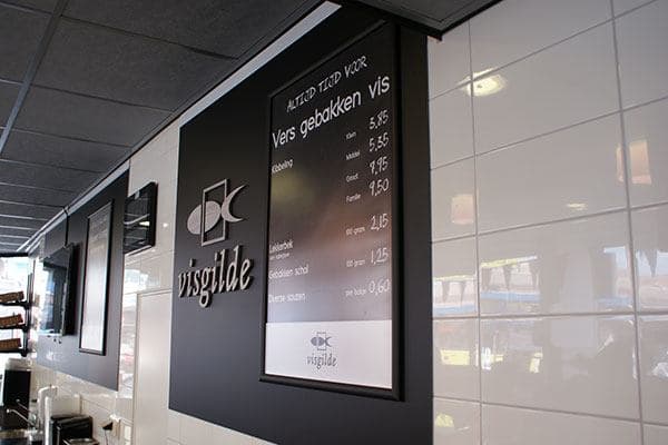 Zwarte wandpanelen met Visgilde-logo en menubord voor gebakken vis, gemonteerd op een wit betegelde winkelwand in een moderne viszaak.