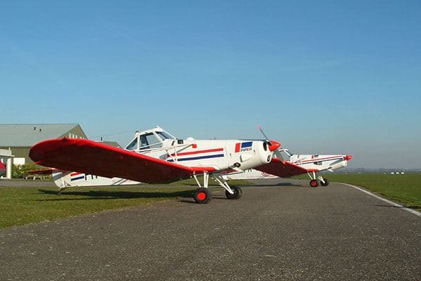 Twee wit-rode sportvliegtuigen met Piper-logo, CNE air-belettering en rode striping op de romp, geparkeerd op een taxibaan naast een hangar.