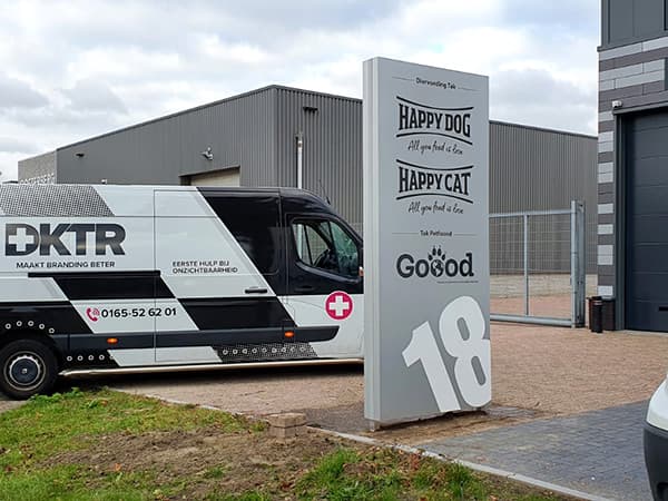 Vrijstaande reclametotem met merkbelettering voor diervoeding naast een zwart-wit bestickerde bedrijfsbus op een bedrijventerrein.