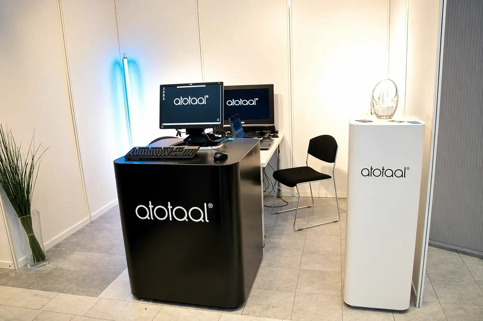 Minimalistische presentatiehoek met zwarte balie en witte displayzuil met atotaal-belettering, computerschermen met logo en strakke kantoorinrichting.
