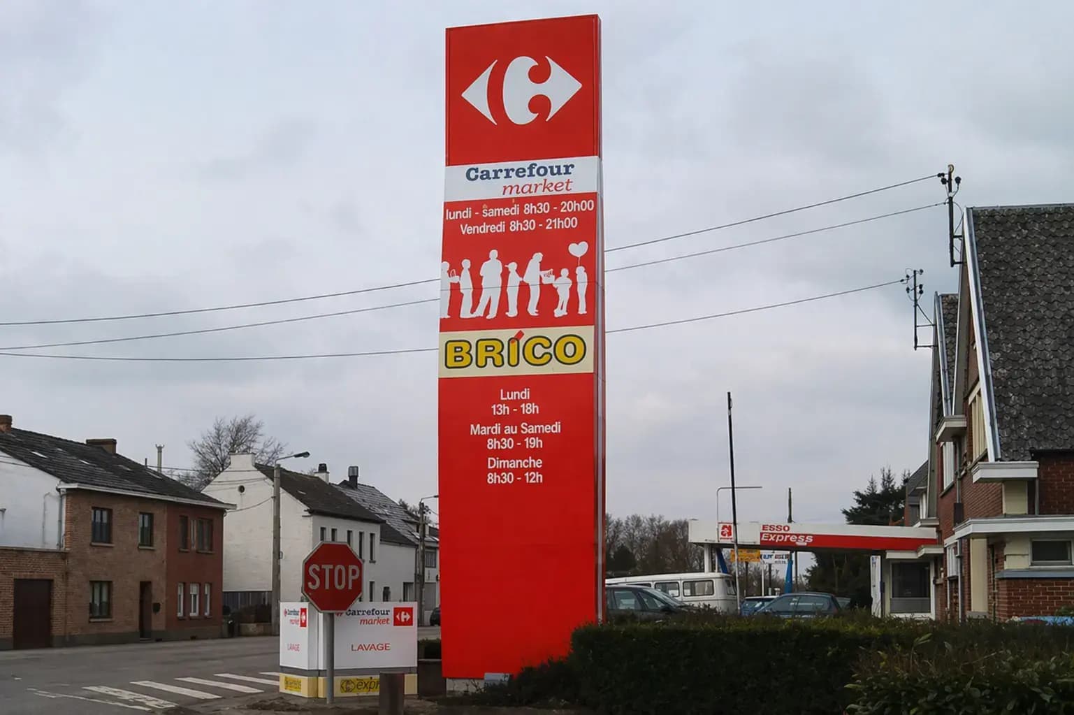 Hoge rode reclamezuil met Carrefour market en BRICO langs een straat bij een winkel- en tankstationlocatie.
