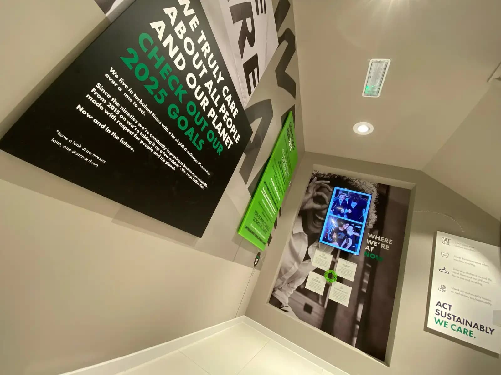 Hoek van een winkelinterieur met grote wandpanelen, interieurbelettering en digitale display over duurzaamheid, waaronder de teksten "CHECK OUT OUR 2025 GOALS", "WHERE WE’RE AT NOW" en "ACT SUSTAINABLY WE CARE."