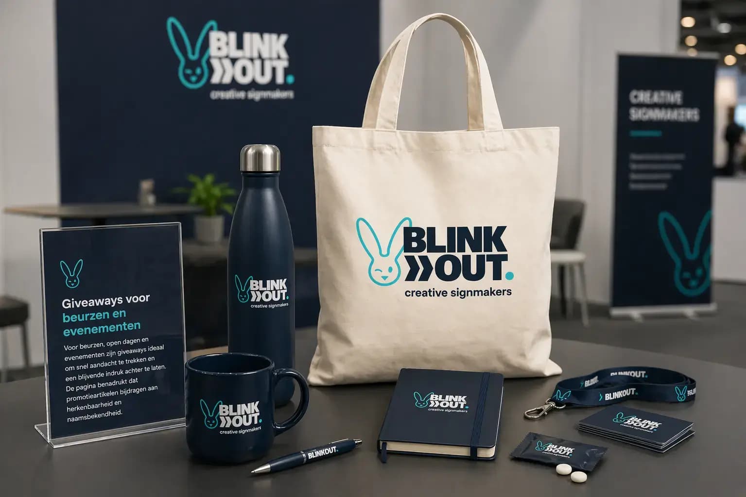 Presentatie van promotionele relatiegeschenken met BLINK OUT creative signmakers-logo, waaronder een draagtas, thermosfles, mok, notitieboek, pen, keycord en flyers op een beursstand.