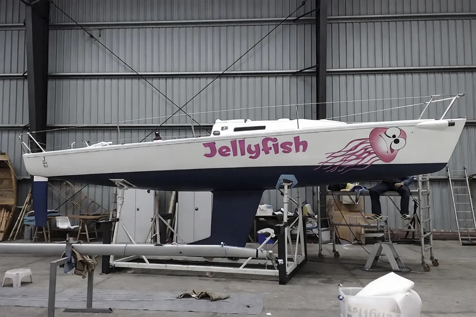 Witte zeilboot op bok in een werkplaats met roze bootbelettering ‘Jellyfish’ en een grote kwalillustratie op de romp.