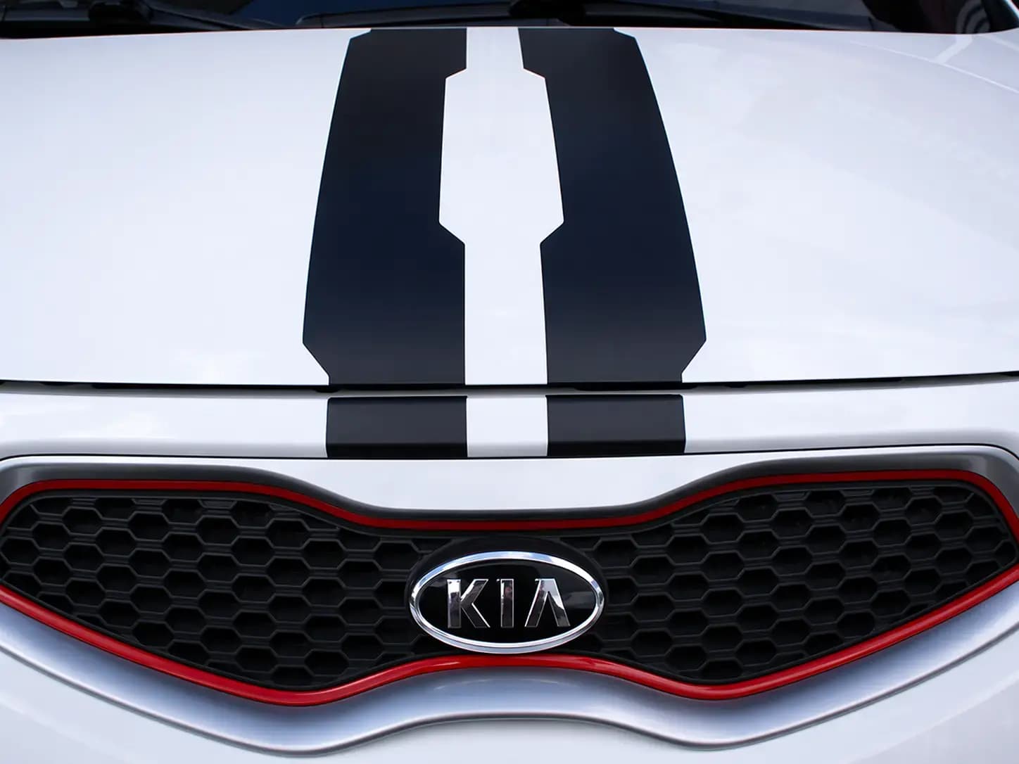 Close-up van de voorzijde van een witte Kia met zwarte striping op de motorkap en rode accenten rond de grille, geschikt als voorbeeld van voertuigbelettering en car wrapping.