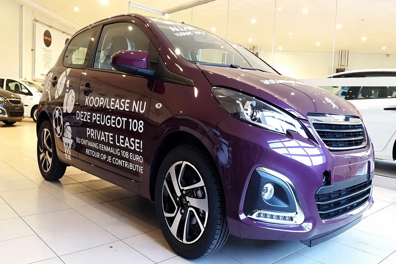 Paarse Peugeot 108 met witte voertuigbelettering en promotionele autobelettering op de zijkant, gefotografeerd in een showroom met duidelijke reclameletters en vinylstickers.