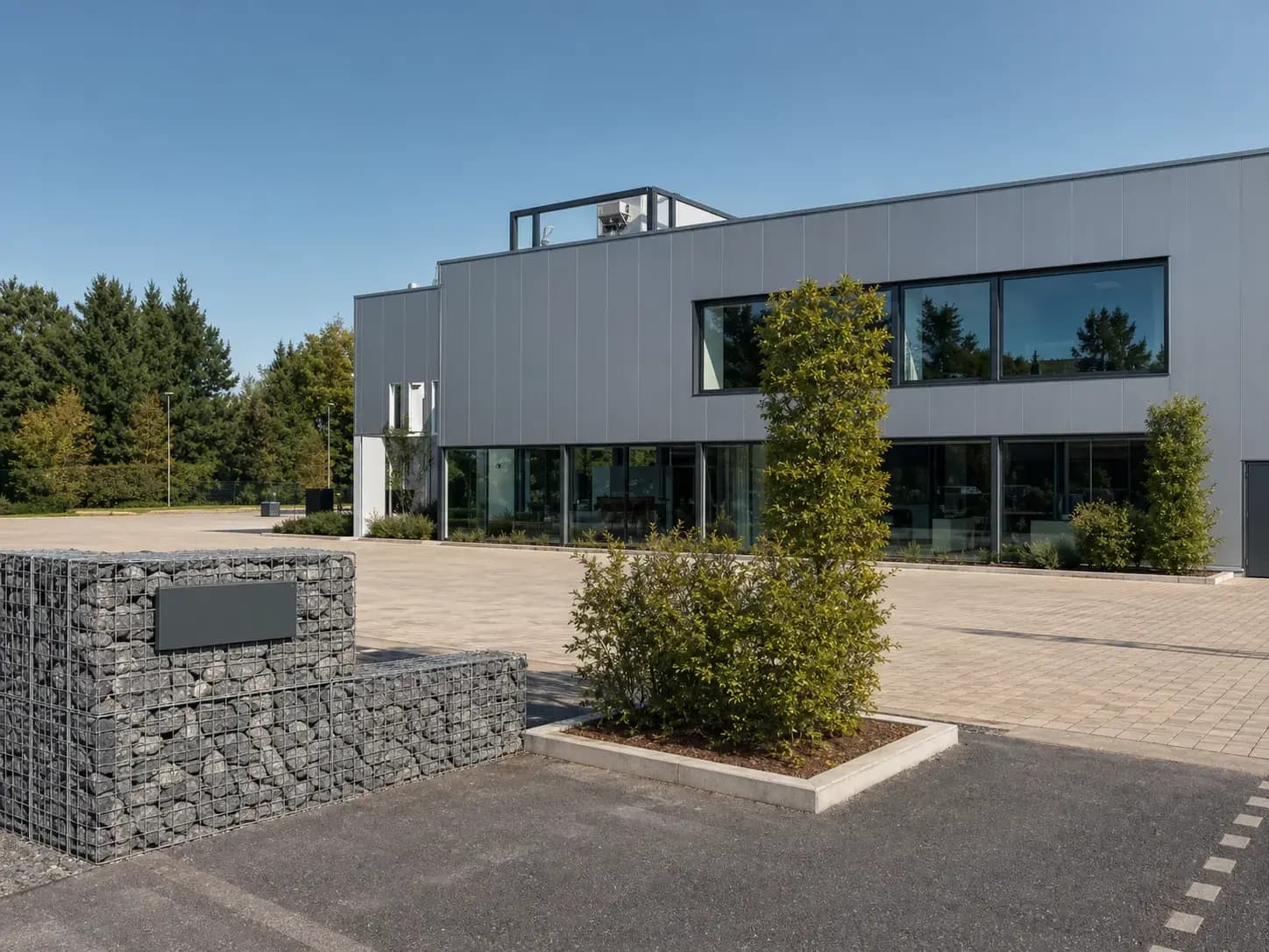 Modern bedrijfspand met grijze gevelbekleding, grote glaspartijen en een rechthoekig gevelbord op een schanskorf bij de entree.