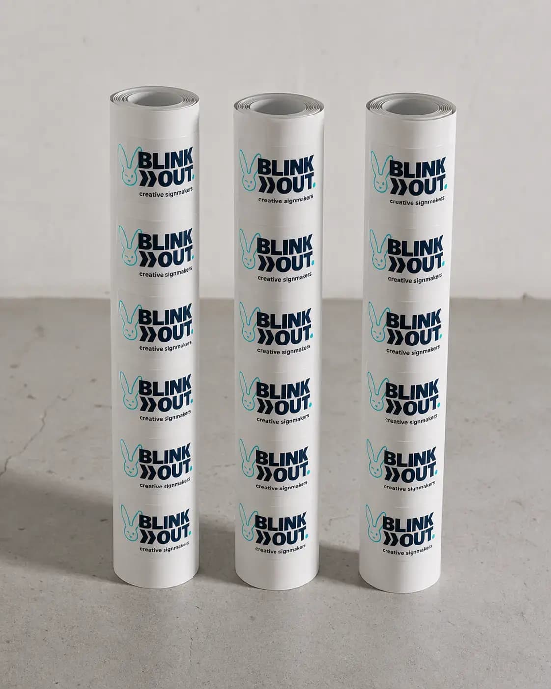 Drie rollen witte etiketten met herhaald BLINK OUT creative signmakers-logo, gepresenteerd als bedrukte stickers op een neutrale ondergrond.