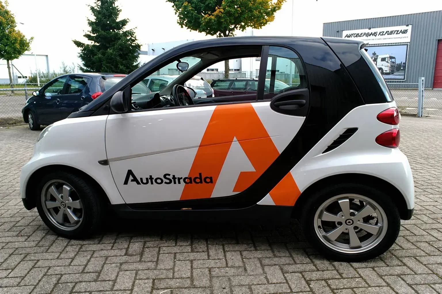Zijaanzicht van een compacte witte stadsauto met zwarte panelen en opvallende oranje AutoStrada voertuigbelettering op de deur en achterzijde, geparkeerd op een bedrijventerrein.