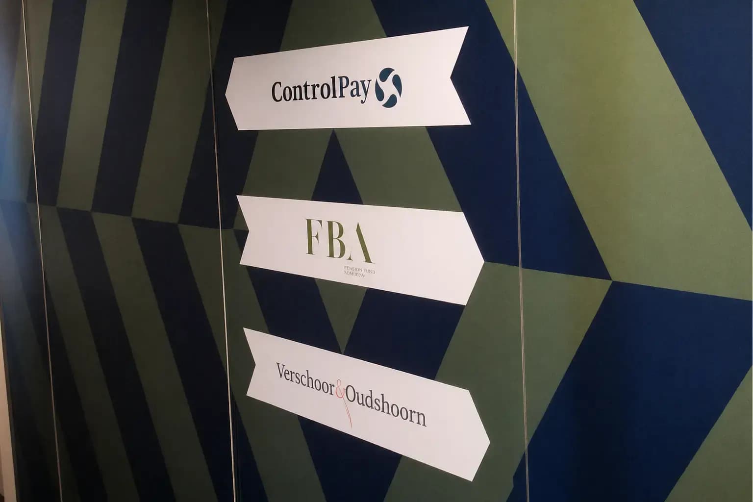 Wayfinding-wand met geometrische wandbekleding en witte richtingsborden voor ControlPay, FBA en Verschoor & Oudshoorn als kantoorbewegwijzering.