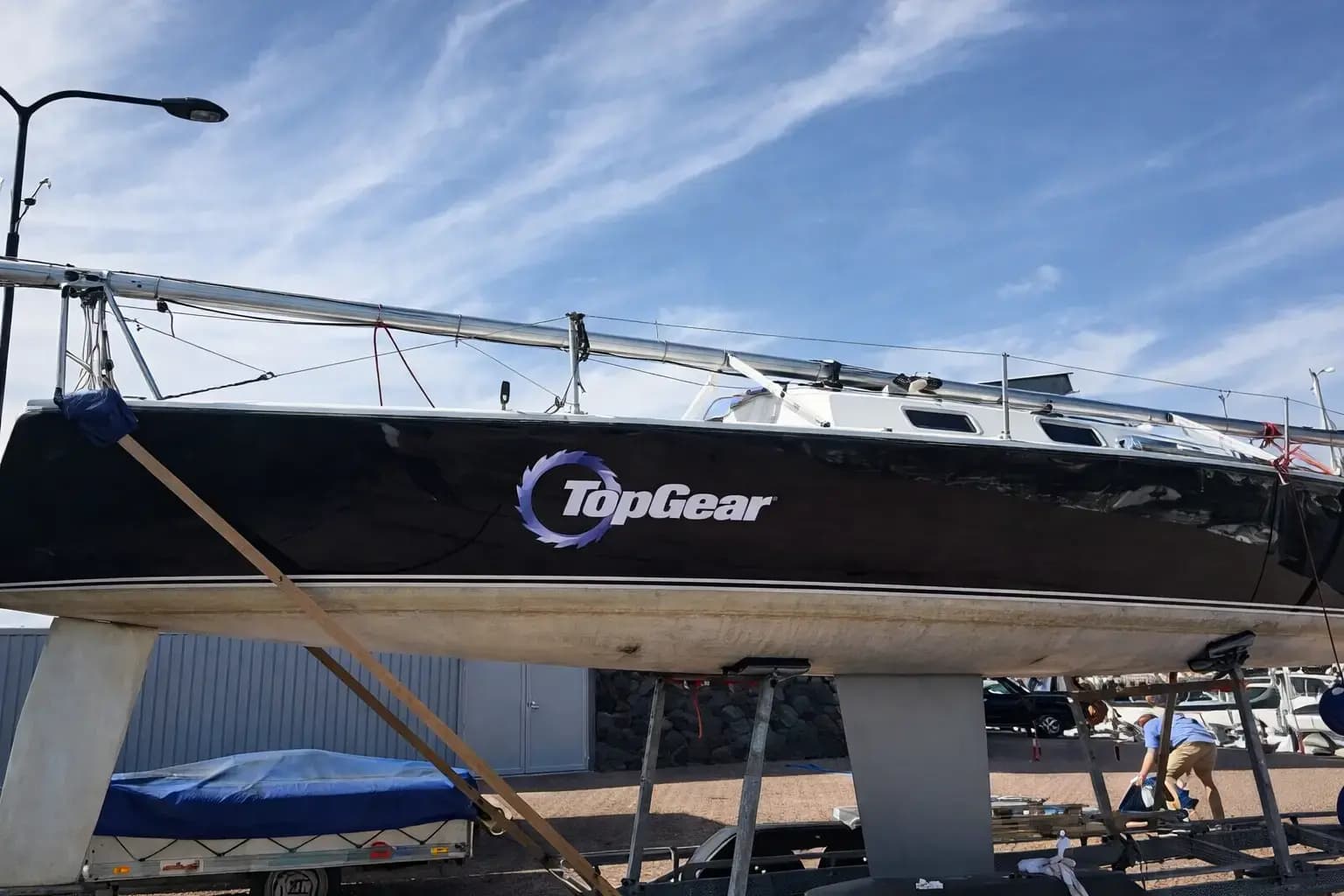 Zwarte zeilboot op bok met grote TopGear-belettering en ronde logosticker op de romp, gefotografeerd in een jachthaven onder een heldere lucht.
