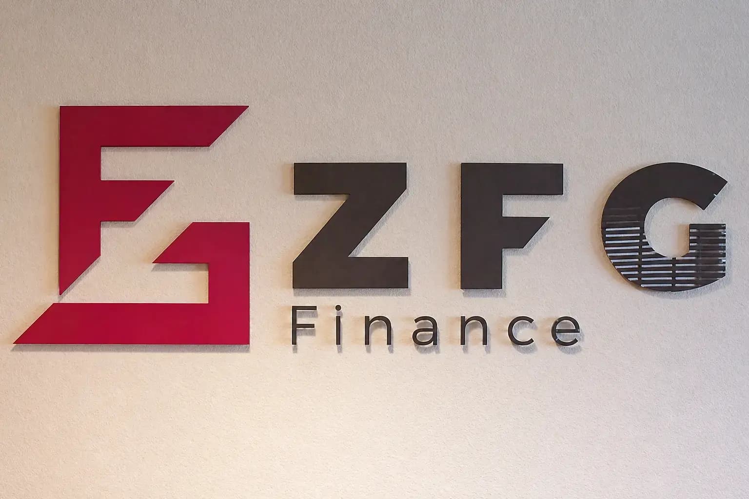 ZFG Finance wandlogo met rode beeldmerkplaat en zwarte 3D letters, gemonteerd op een lichte structuurmuur als zakelijke binnenreclame.