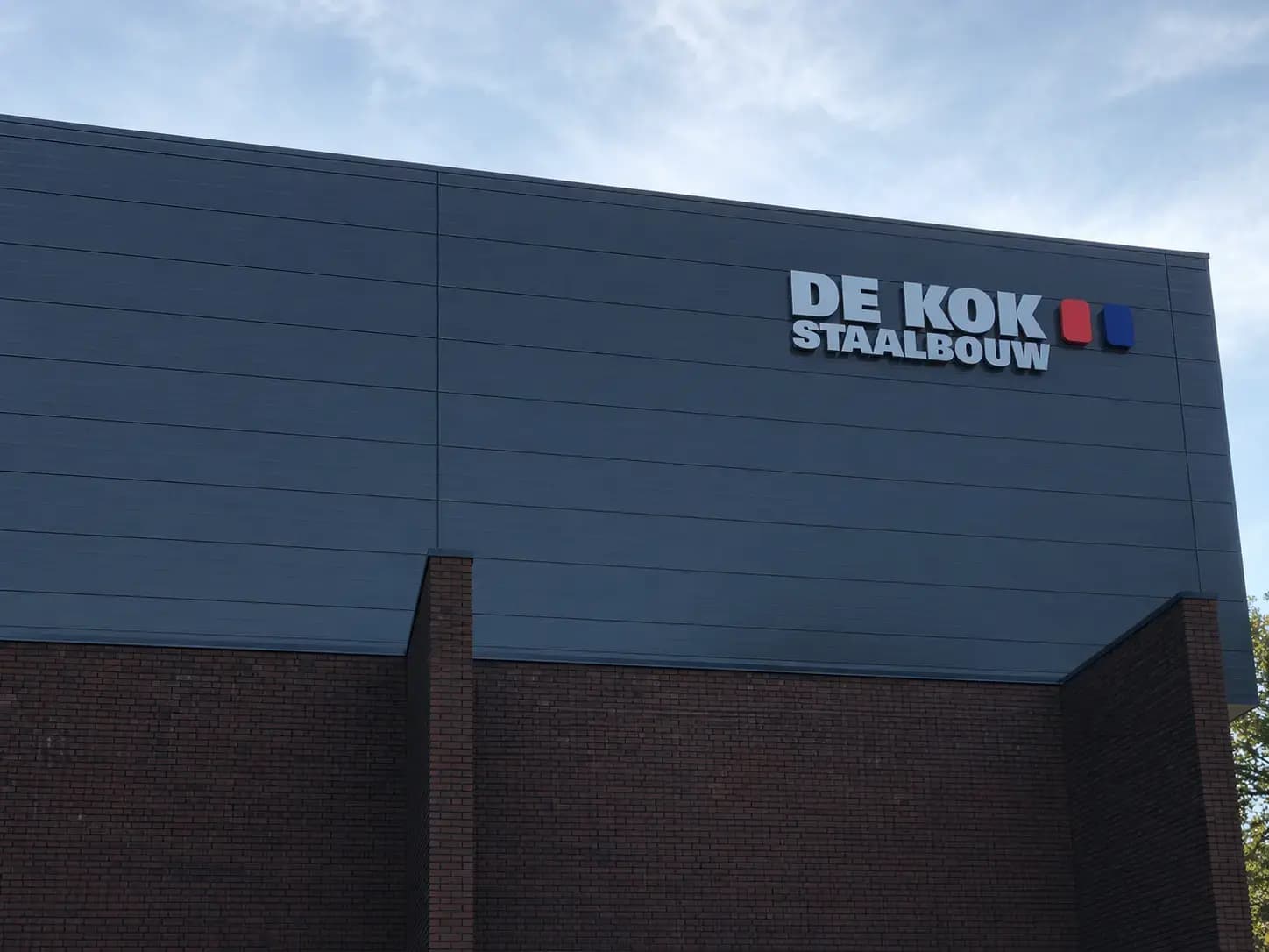 Moderne bedrijfshal met gevelreclame van DE KOK STAALBOUW in witte 3D-letters en rood-blauw logo op donkergrijze gevelpanelen boven een bakstenen plint.