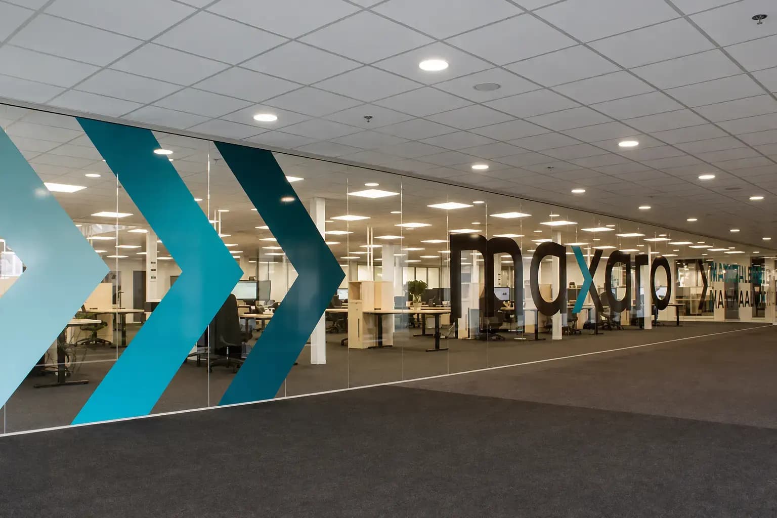 Glaswandbelettering met turquoise pijlen en groot bedrijfslogo op een glazen kantoorwand in een open kantoorruimte.