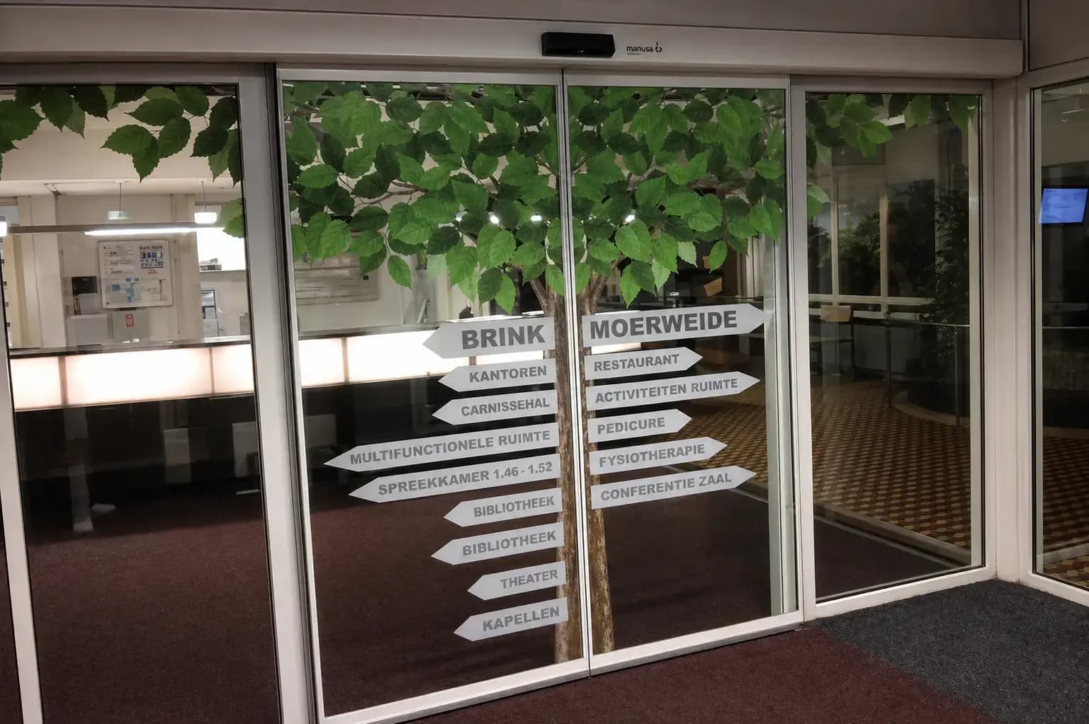 Glasdeuren met raamfolie van een boom en grijze wayfinding-pijlen voor Brink, Moerweide, kantoren en voorzieningen bij een entree.