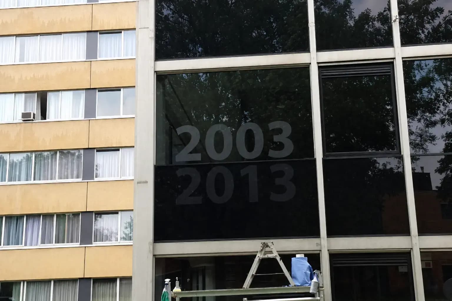 Donkere raamfolie met uitgespaarde jaartallen 2003 en 2013 op een glazen gevel, aangebracht boven een werkplatform met ladder.