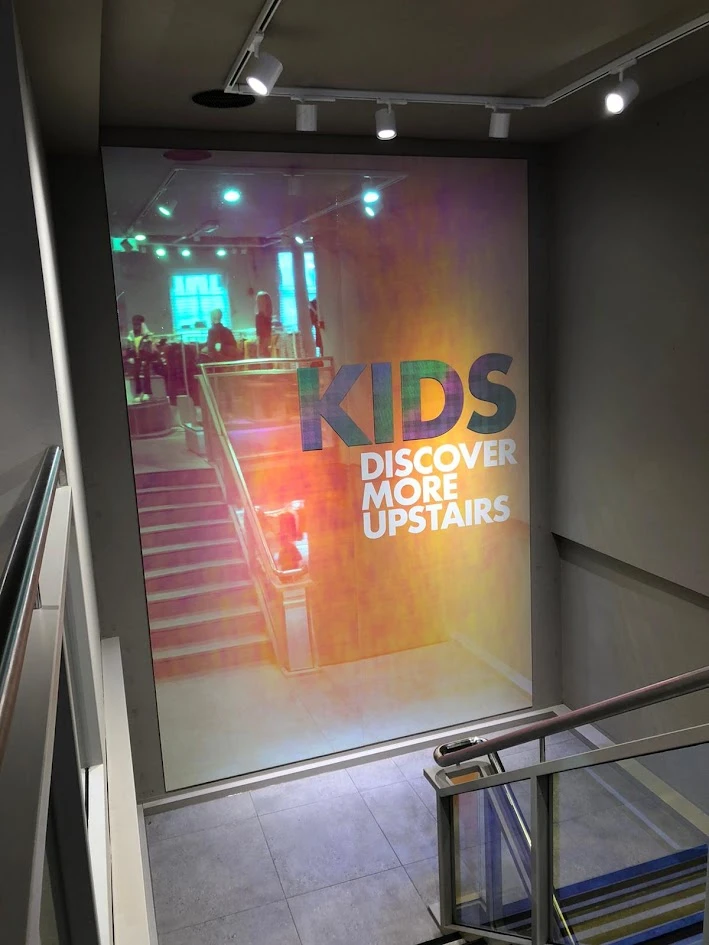 Dichroic folie op een witte Dibond plaat bij een winkeltrappenhuis, met de tekst Kids Discover More Upstairs