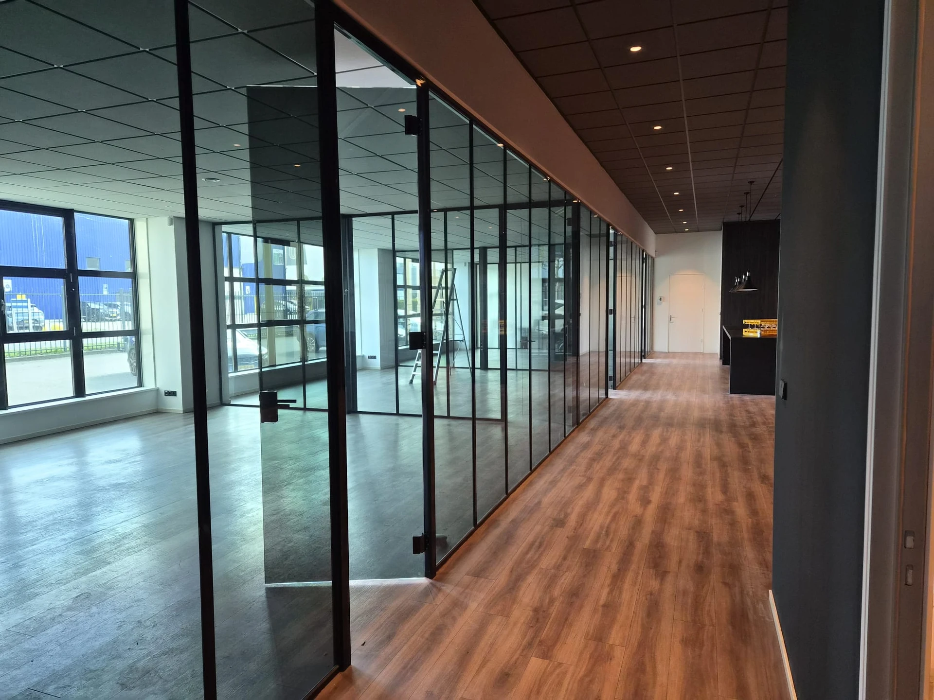 Lange gang met donker getinte glazen kantoorwanden, zwarte profielen en een houten vloer